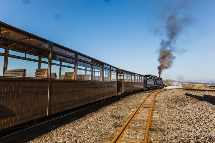 Steam train on curved tracks with wooden passenger cars and smoke stack in clear blue sky.