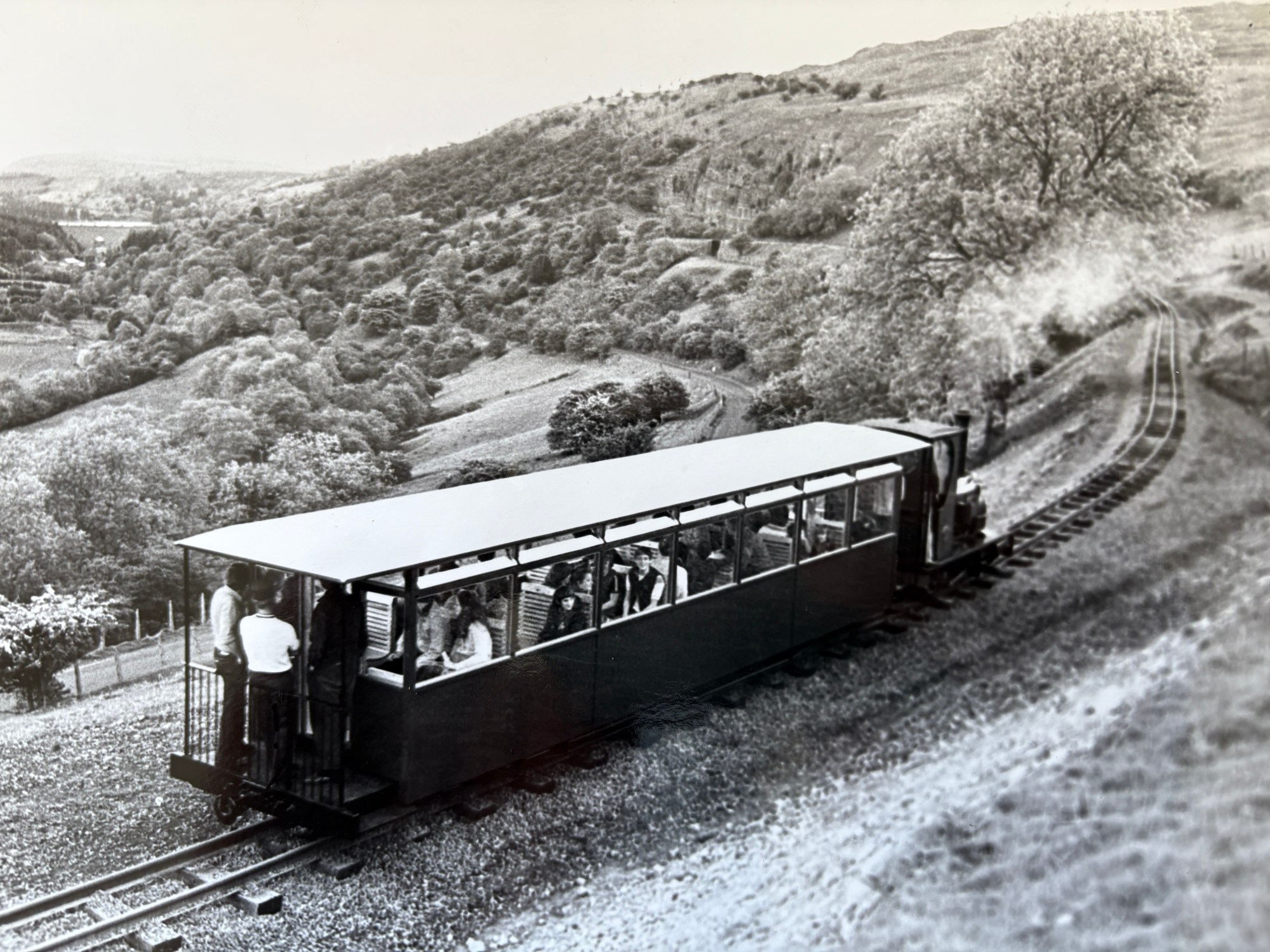 IMG_1638 Scenic hillside train with passengers overlooking a lush, rolling landscape.