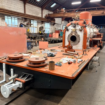 Partially assembled steam locomotive in a workshop with scattered tools on metal platform.