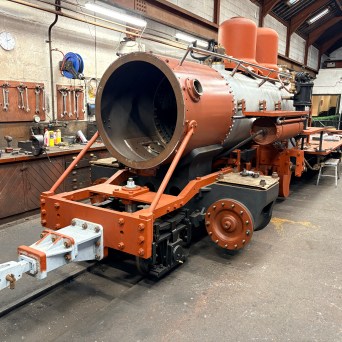 Steam locomotive under restoration in a workshop, with tools and equipment nearby.