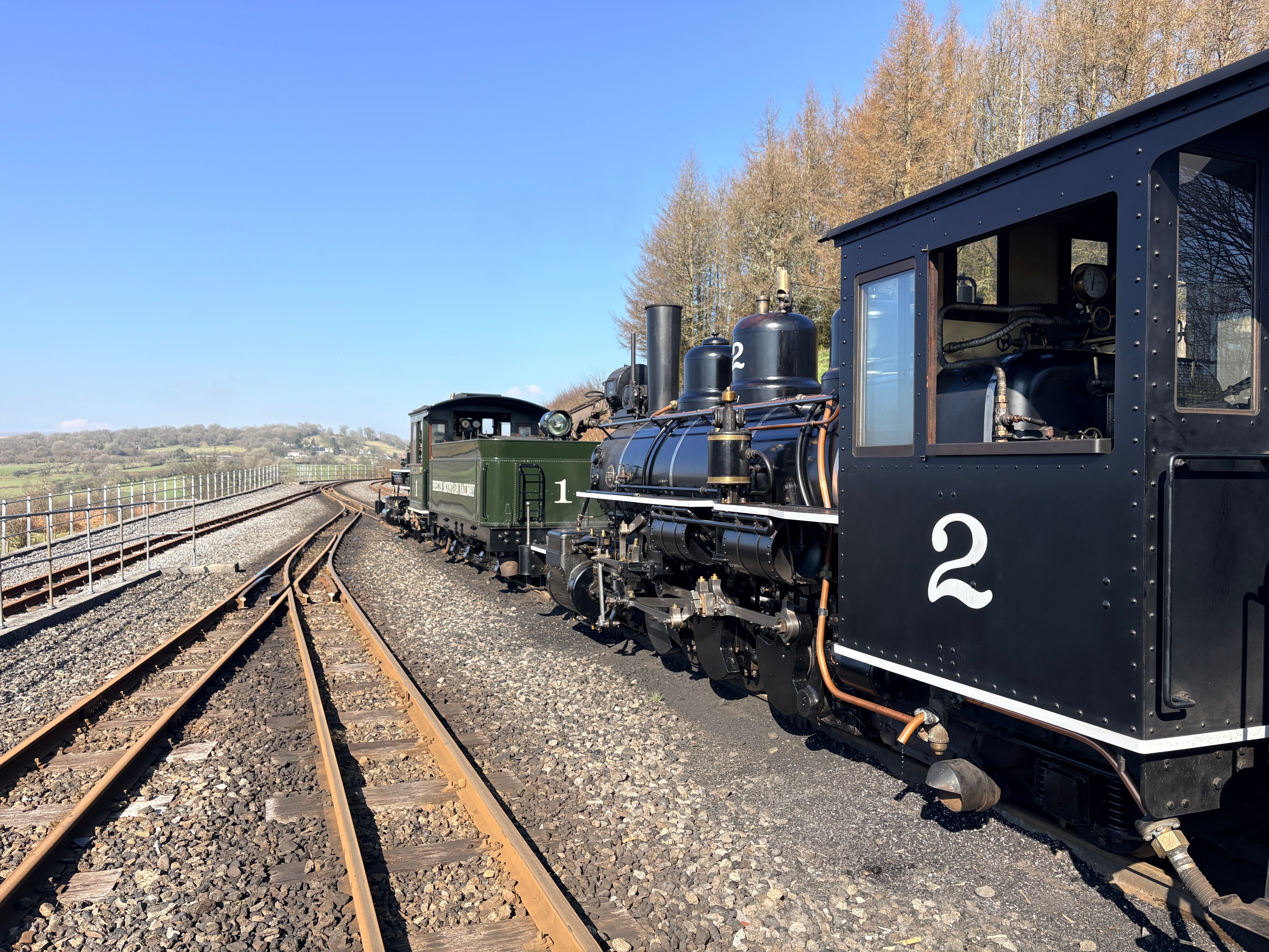 Two vintage steam locomotives on parallel tracks, trees and clear sky in the background.