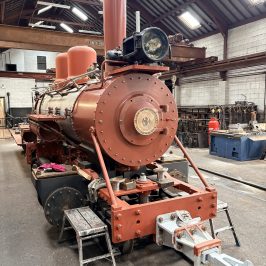Old steam locomotive in a workshop, surrounded by tools and machinery.