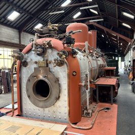 Steam engine machinery inside an industrial building with a high wooden ceiling.