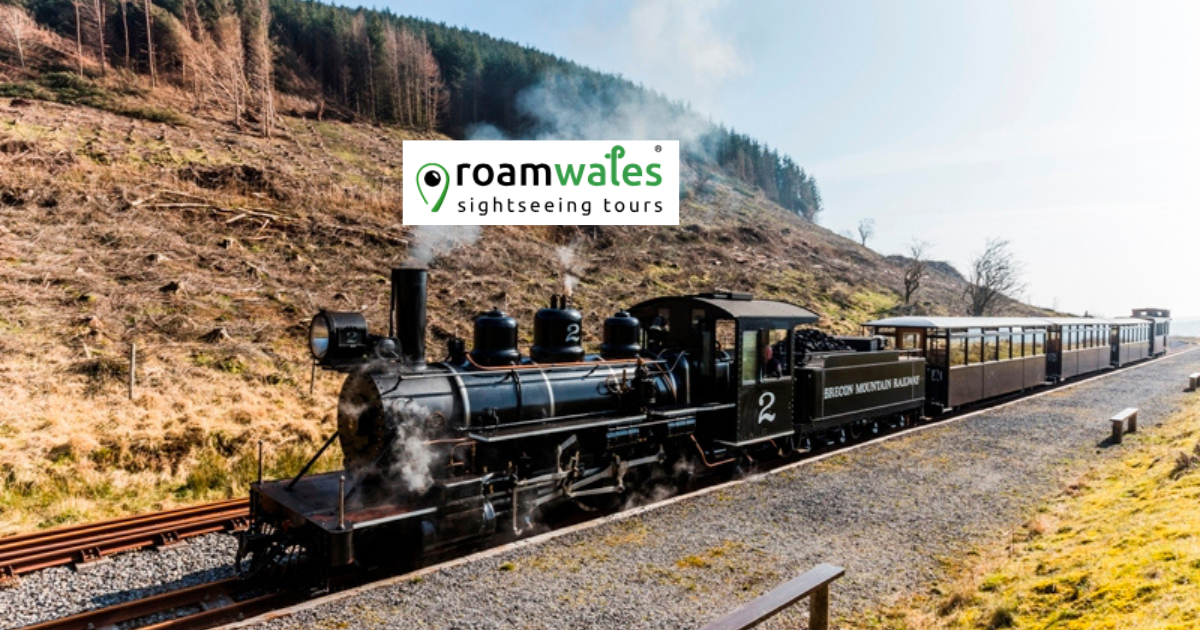 Vintage steam train on rural tracks, hills in background, with 'Roam Wales' logo above.