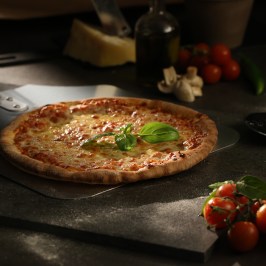 Fresh pizza with basil on stone, surrounded by tomatoes and garlic.