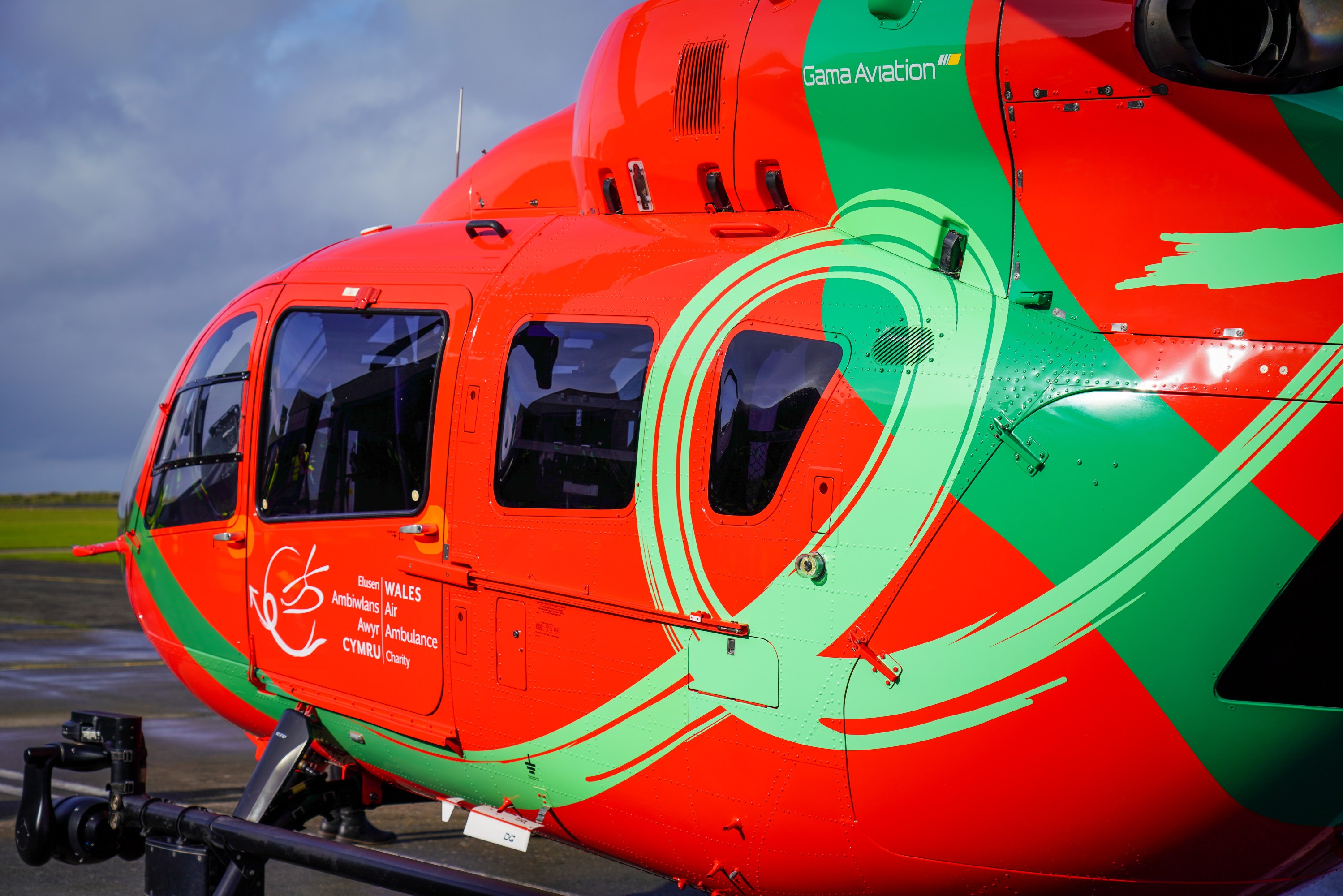 Red and green helicopter with Wales Air Ambulance logo on a tarmac.