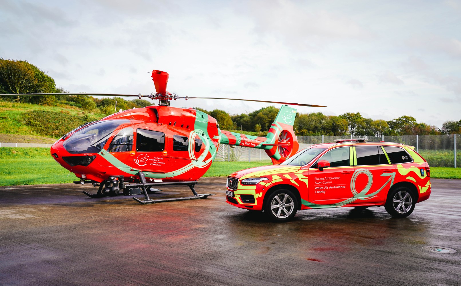Red and green helicopter and SUV on a tarmac.